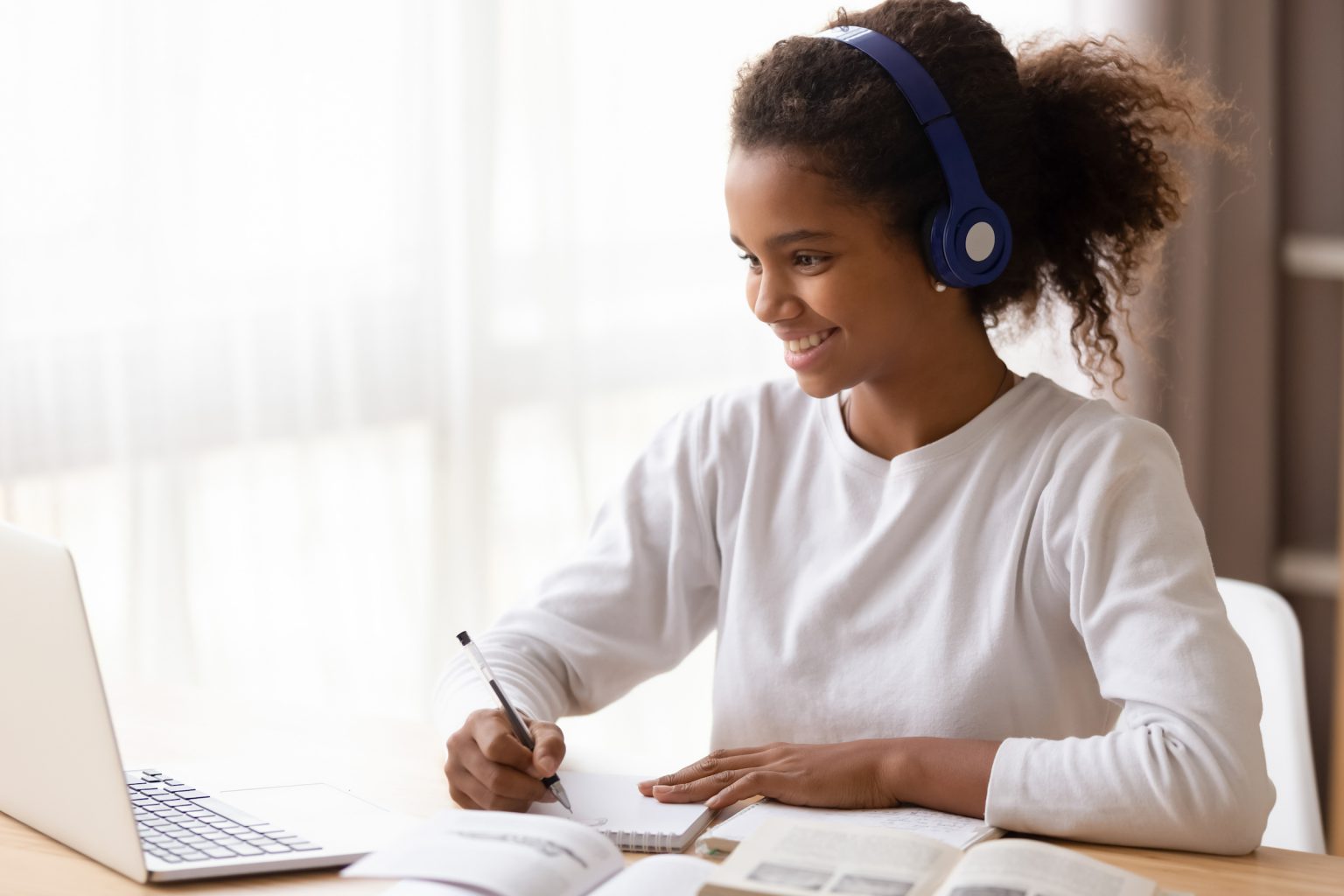 Happy african schoolgirl doing homework sitting at desk, teen girl feels glad studying online e-learning use internet wear headphones writing noting. Education and knowledge modern technology concept